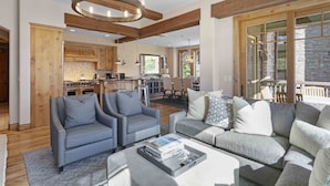A spacious living area with a grey sofa, two armchairs, and a coffee table. A kitchen with wooden cabinets and a stone fireplace is visible in the background. - 302 - Four Bedroom Condo Silver Strike by Deer Valley Resort (Park City)
