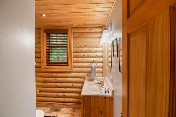 A wooden cabin bathroom with a wooden wall, a window with blinds, a sink with a mirror, and a door. - Er25 - Country Days 2 Bedroom Cabin (Pigeon Forge)