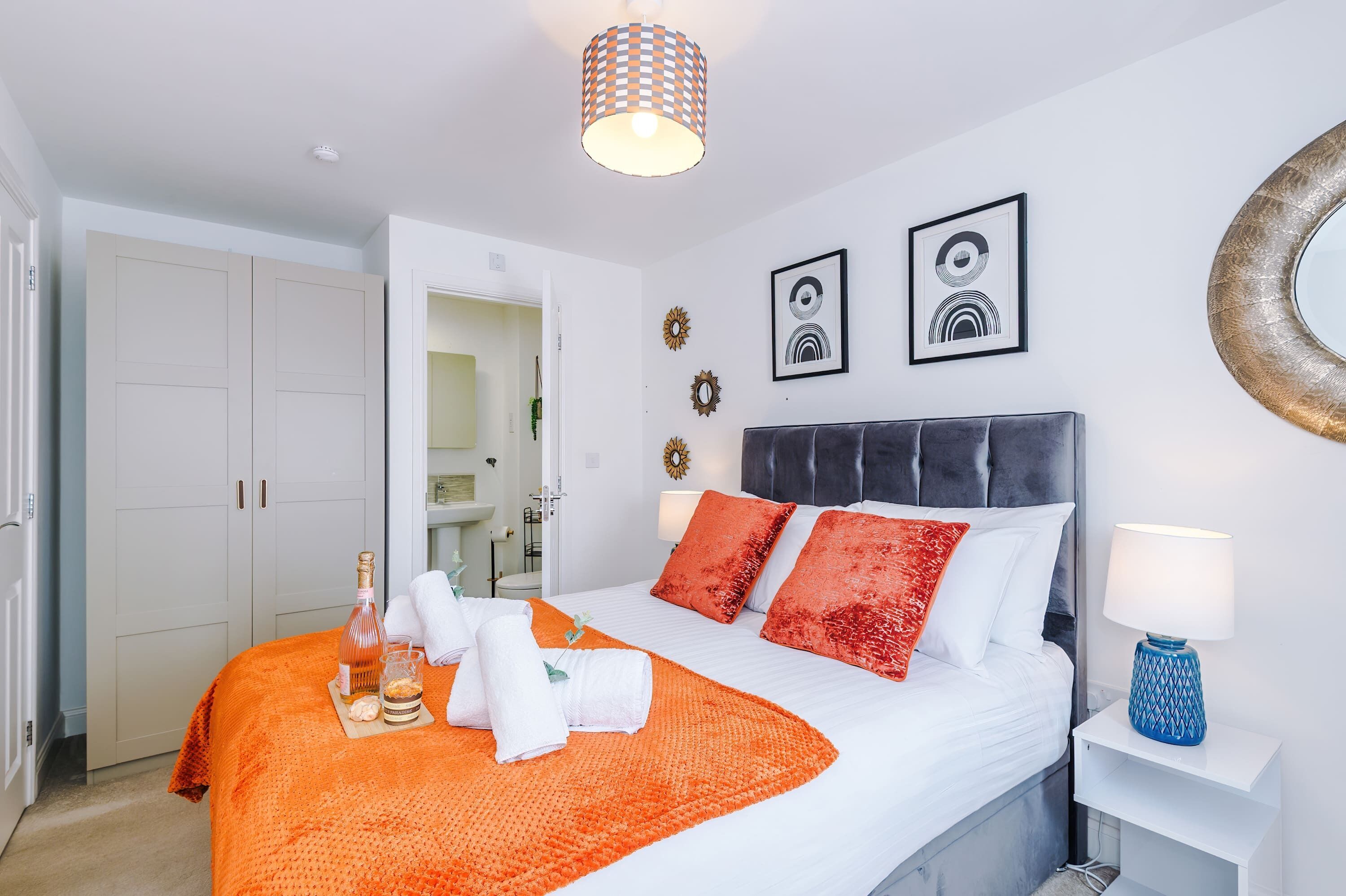 Cama bem arrumada com colcha branca e laranja, duas almofadas vermelhas e uma mesa de cabeceira com abajur azul.