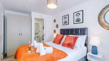Cama bem arrumada com colcha branca e laranja, duas almofadas vermelhas e uma mesa de cabeceira com abajur azul.
