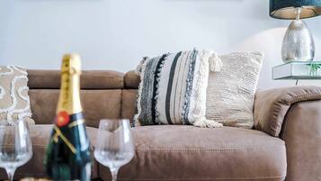 Um sofá de couro marrom com um cobertor listrado e uma almofada com franjas. À frente do sofá, há uma garrafa de champanhe e duas taças de vinho vazias. Uma luminária com cúpula metálica está sobre uma mesinha lateral à direita.