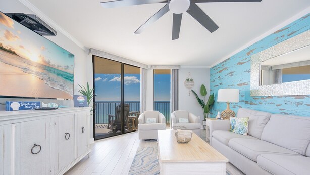 A living room with a ceiling fan, a large painting of a beach scene, a sofa, a coffee table, and a view through the glass doors. - Phoenix V 1009 2 Bedroom Condo (Orange Beach)
