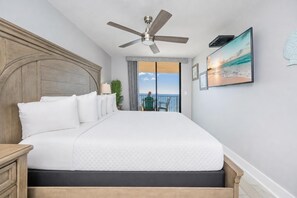 Une chambre avec un grand lit, un ventilateur de plafond, une vue sur l’océan par une porte coulissante et un tableau encadré au mur.