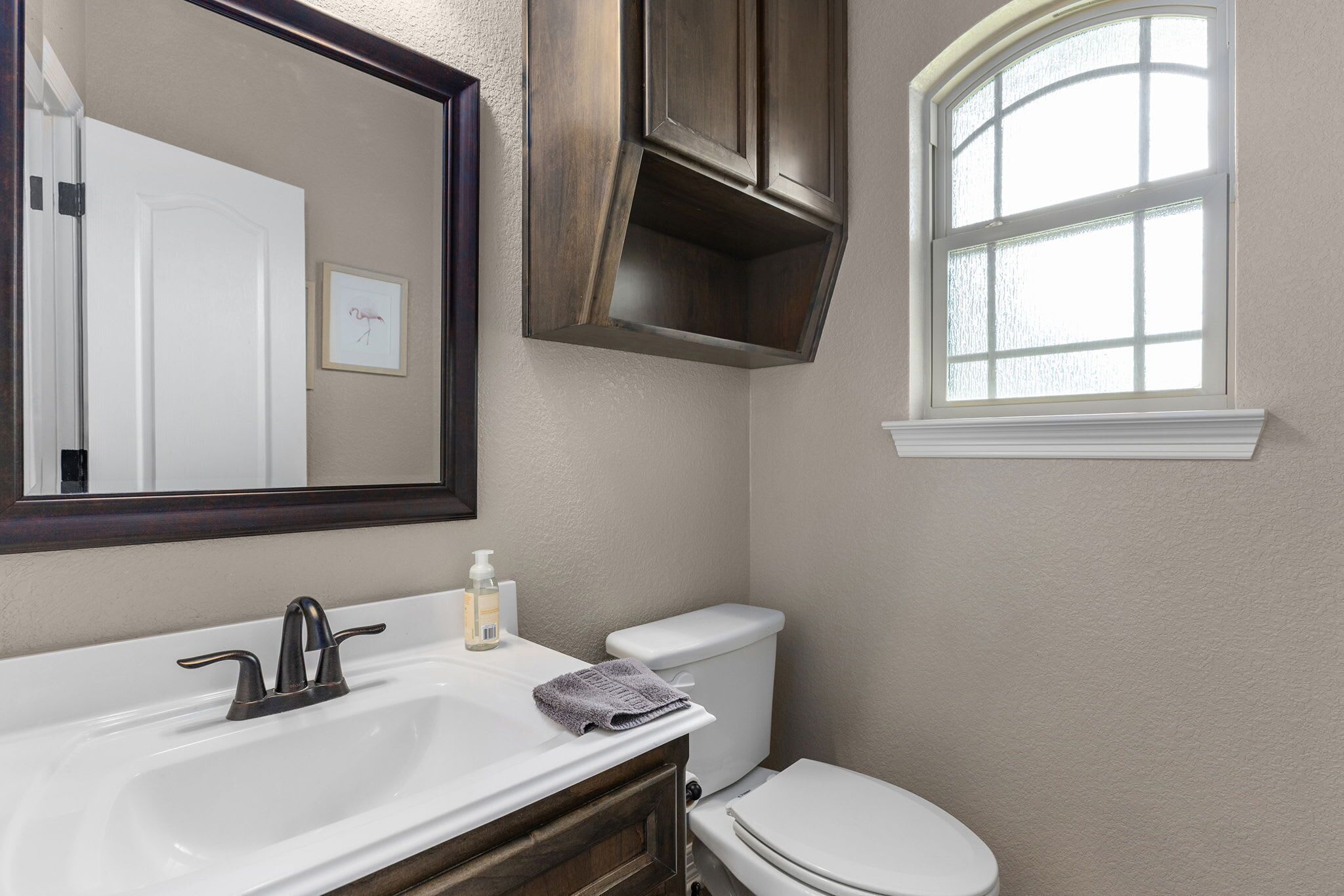 Un baño con lavamanos blanco, mueble oscuro, inodoro blanco y ventana con vidrio satinado.