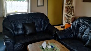 A black leather sofa, a wooden coffee table with a green placemat, and a patterned curtain. - Apartment 'Fewo Nordstern Familie Sauerwald' with Shared Garden and Wi-Fi (Pellworm)