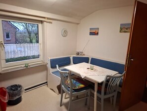 Une petite pièce avec une table et des chaises, une fenêtre donnant sur un immeuble en briques, une horloge et une radio.