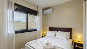 A hotel room with a bed, a window, an air conditioner, and bedside lamps. - Apartment in Condominium With Gym in PoA - Ips0701 (Porto Alegre)