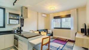 A compact kitchen with a microwave, stove, and modern cabinetry. There is a dining area with a table and chairs, and a sofa in the living area. A large window provides a view of the cityscape. - Apartment in Condominium With Gym in PoA - Ips0701 (Porto Alegre)