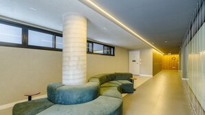 A modern interior with a curved seating area, a tall pillar, and a long hallway with lockers. - Apartment in Condominium With Gym in PoA - Ips0701 (Porto Alegre)