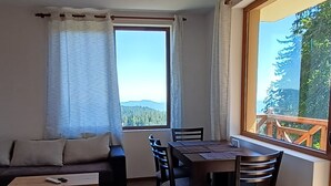 Een kamer met houten vloer, een bank, een eettafel met stoelen en een raam met uitzicht op bomen en bergen.