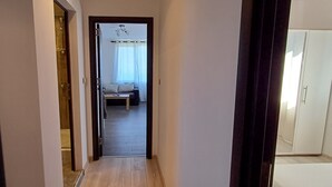 Een smalle gang die leidt naar een slaapkamer met een bed, nachtkastje en een raam met gordijnen.
