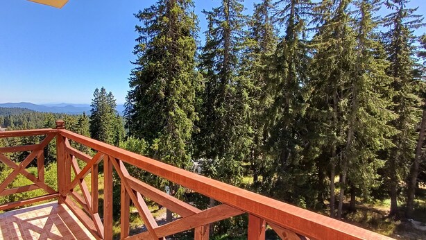 Een balkon met houten hekwerk met uitzicht op een bebost gebied en verre bergen.