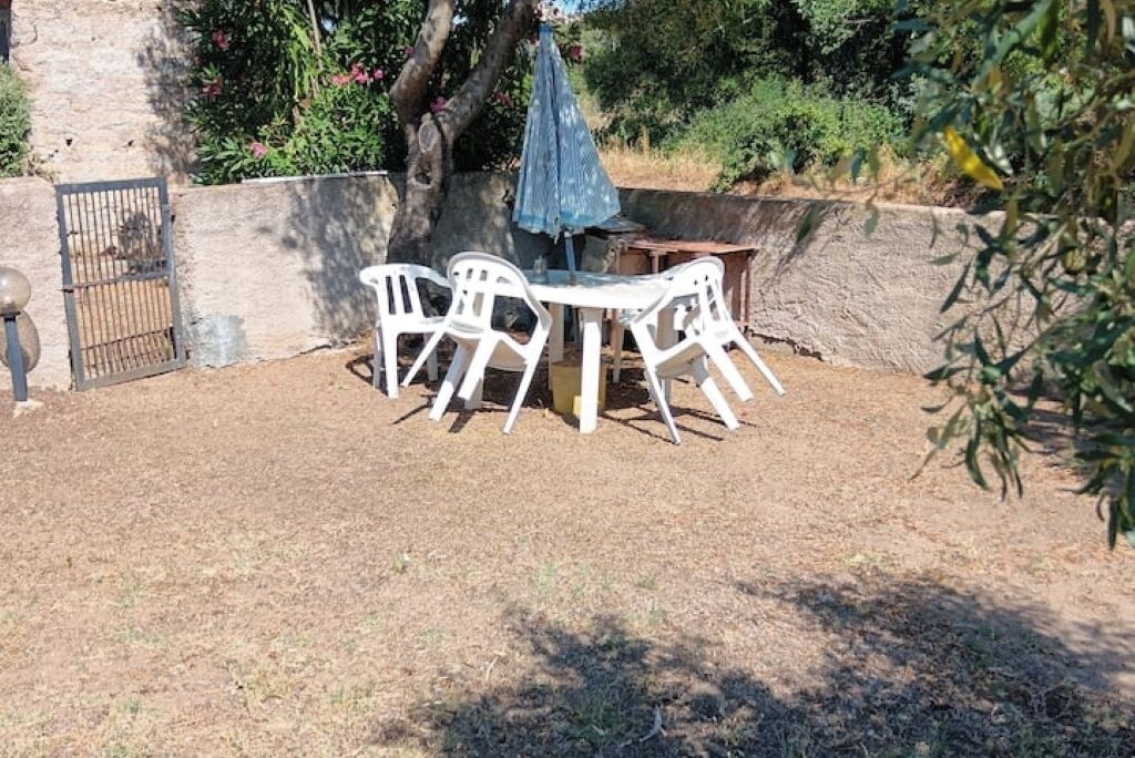 Área externa com assentos, cadeiras de plástico branco e uma mesa, cercada por uma parede de pedra e vegetação.