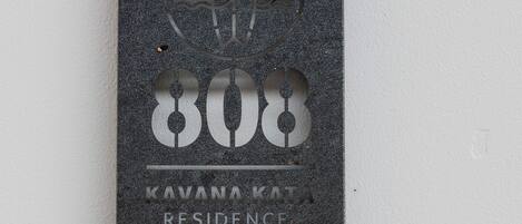 Une plaque sombre, à la texture marquée, ornée d’un emblème circulaire, du chiffre « 808 » et de l’inscription « KAVANA KATA RESIDENCE ».