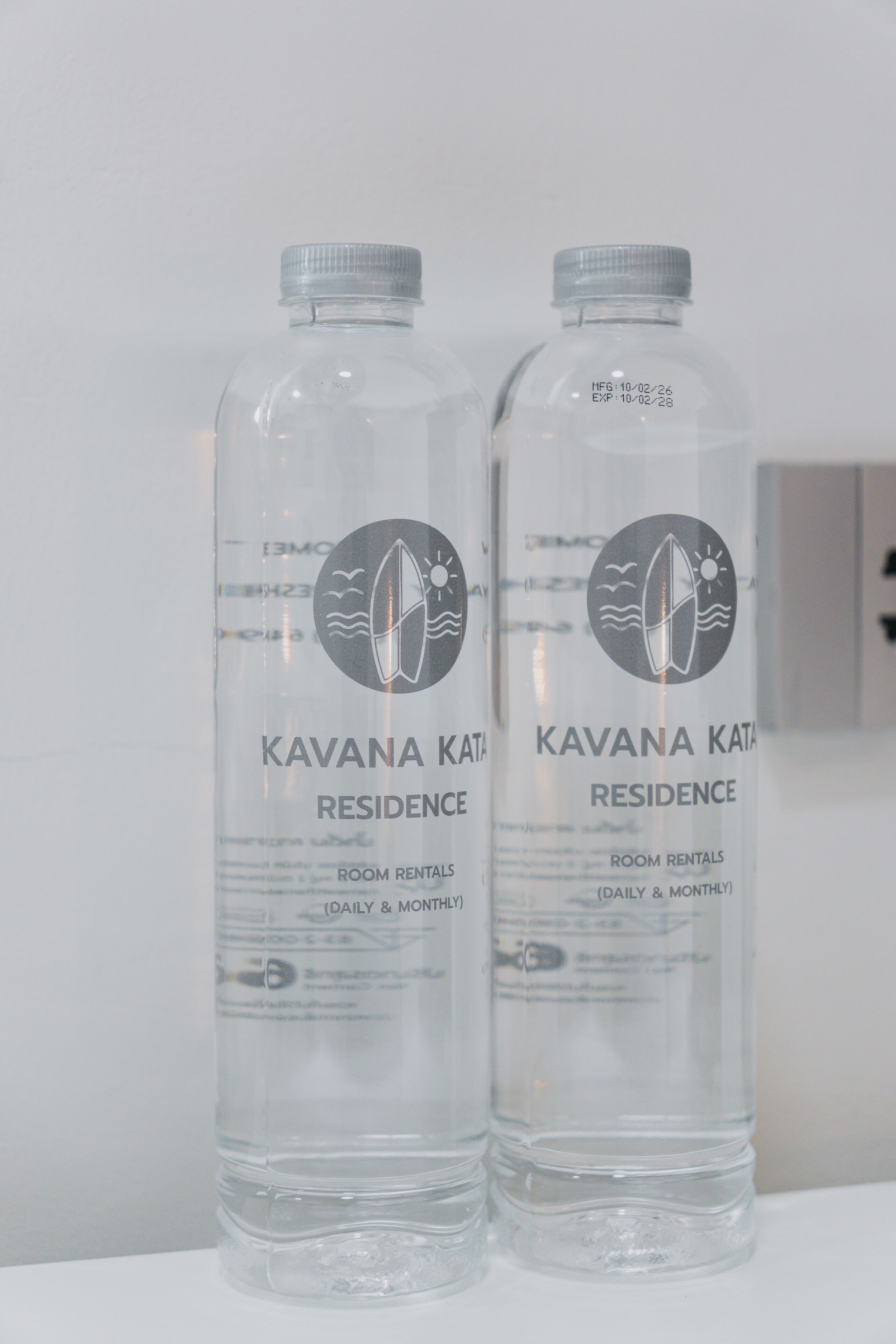 Duas garrafas com o rótulo 'Kavana Kata Residence', apresentando um logotipo com um prancha de surf e ondas.
