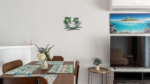 Et spiseområde med et træbord, stole og et lille sofabord med en vase. En vægdekoration viser to fugle på en gren, og et maleri af en tropisk ø.