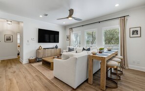Uma sala de estar moderna com sofá, mesa de centro e área de jantar. Há uma TV na parede, um ventilador de teto e janelas grandes com persianas.