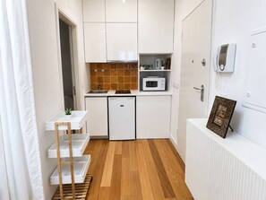 Een compacte keuken met witte kasten, een magnetron, een kleine koelkast en een houten plank.