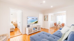 A spacious living room with a flat-screen TV, a blue sofa, and a dining area with a table and chairs. - The Salty Sailor, 7 Market St - Wi-Fi, Water views, Air Con, Boat Parking (Fingal Bay)