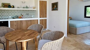 A modern kitchen with a wooden dining table and wicker chairs, a built-in bench, and a view of a bedroom with a window.