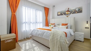 Une chambre à coucher moderne avec un grand lit, du linge de lit blanc, des rideaux orange et une œuvre d’art géométrique accrochée au mur.