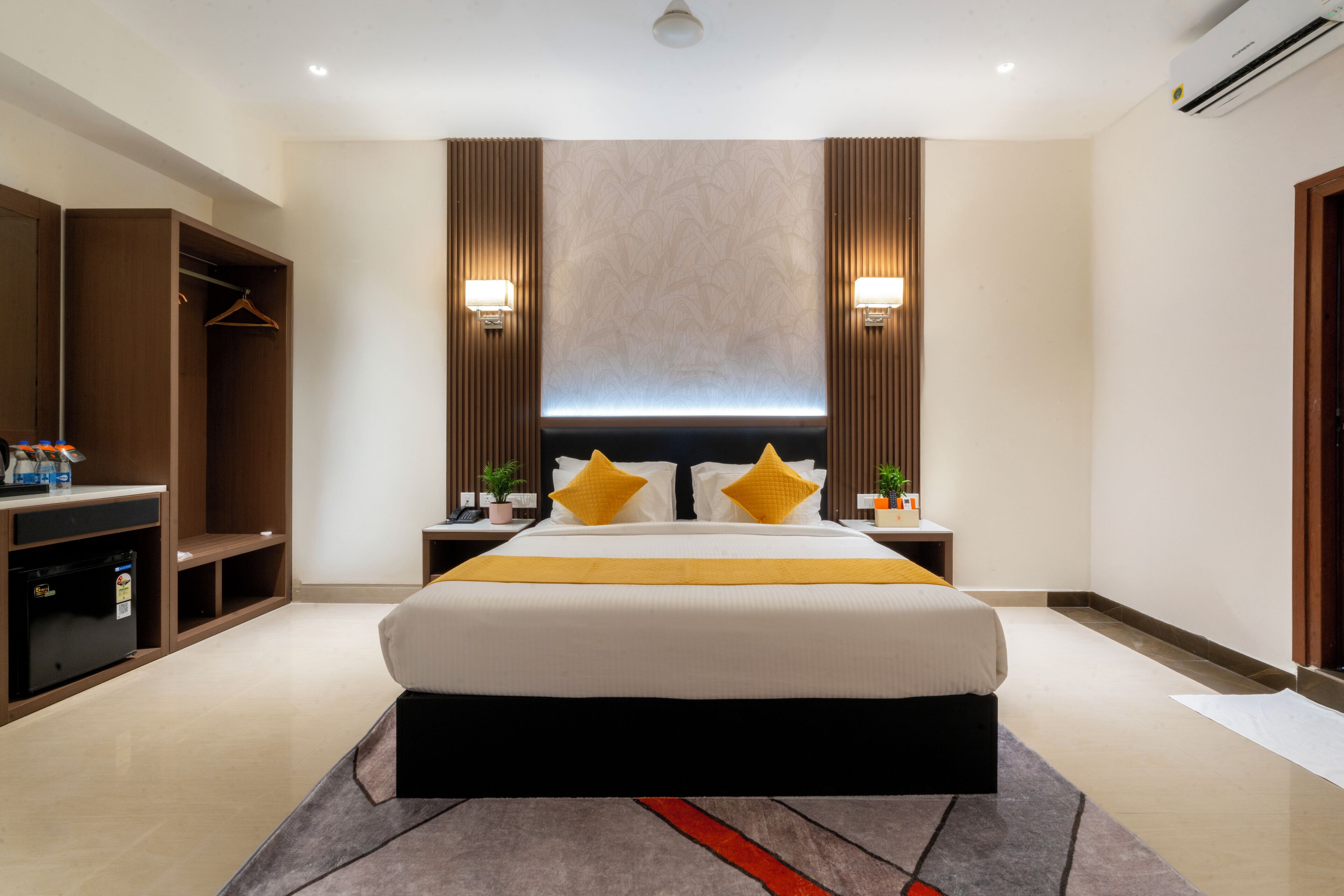 A modern hotel room with a large bed, a wardrobe, a bedside table with a lamp, and a wall-mounted air conditioner.
