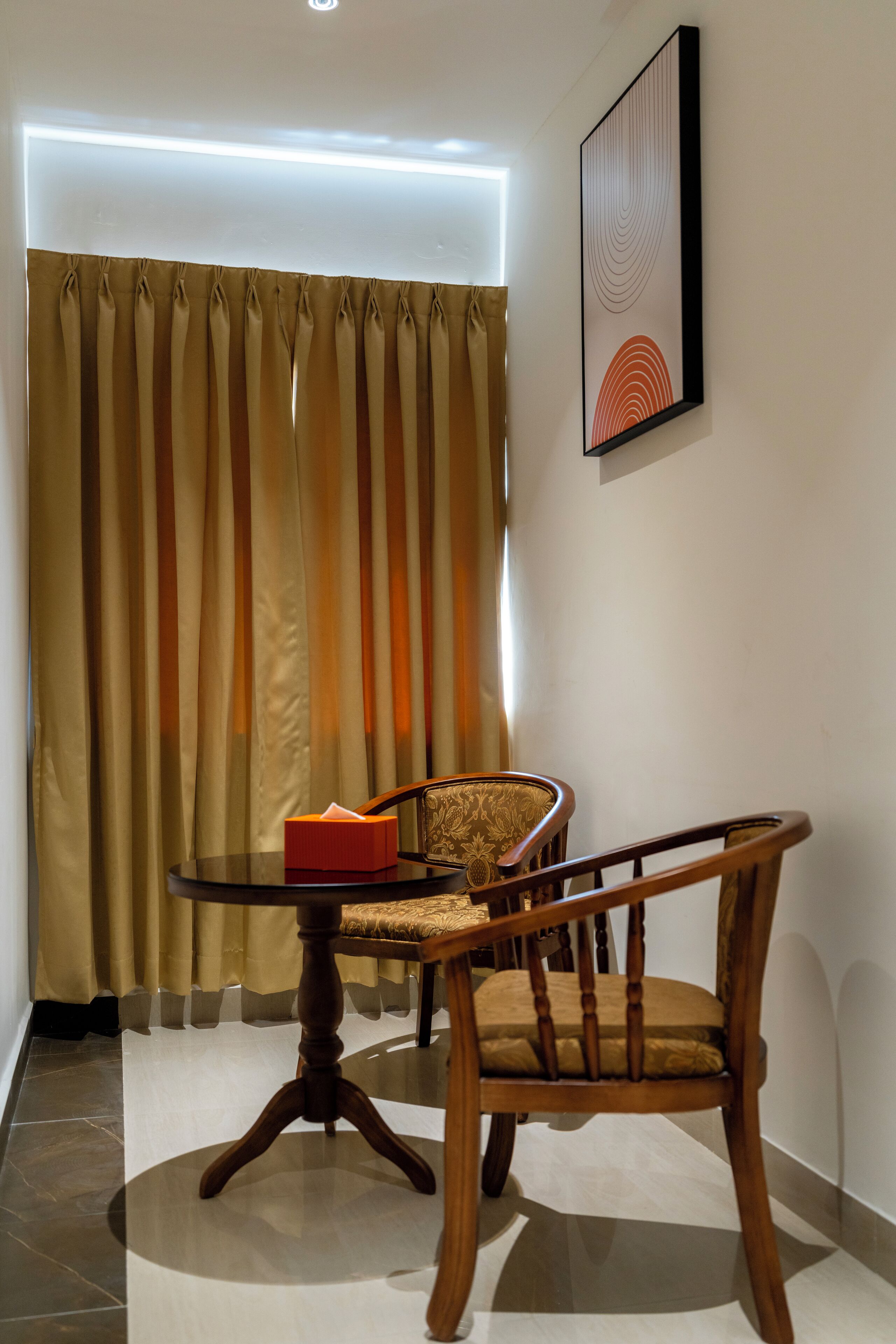 A room with a round wooden table, a chair, a red box, and a framed abstract artwork on the wall.