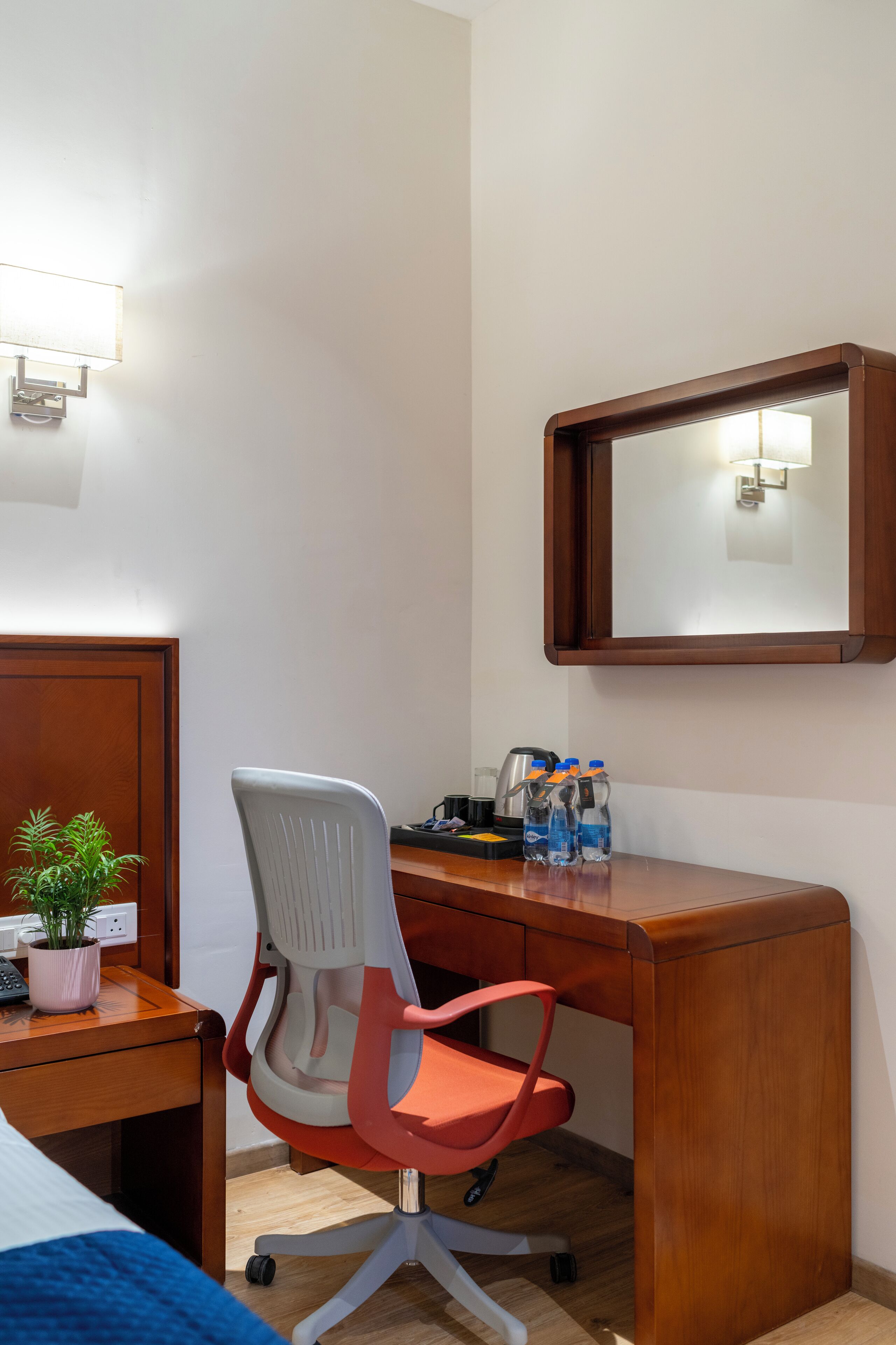 Une chambre d’hôtel avec un bureau en bois, un fauteuil de bureau rouge, une plante en pot, un miroir et une table de chevet avec une lampe et des bouteilles.