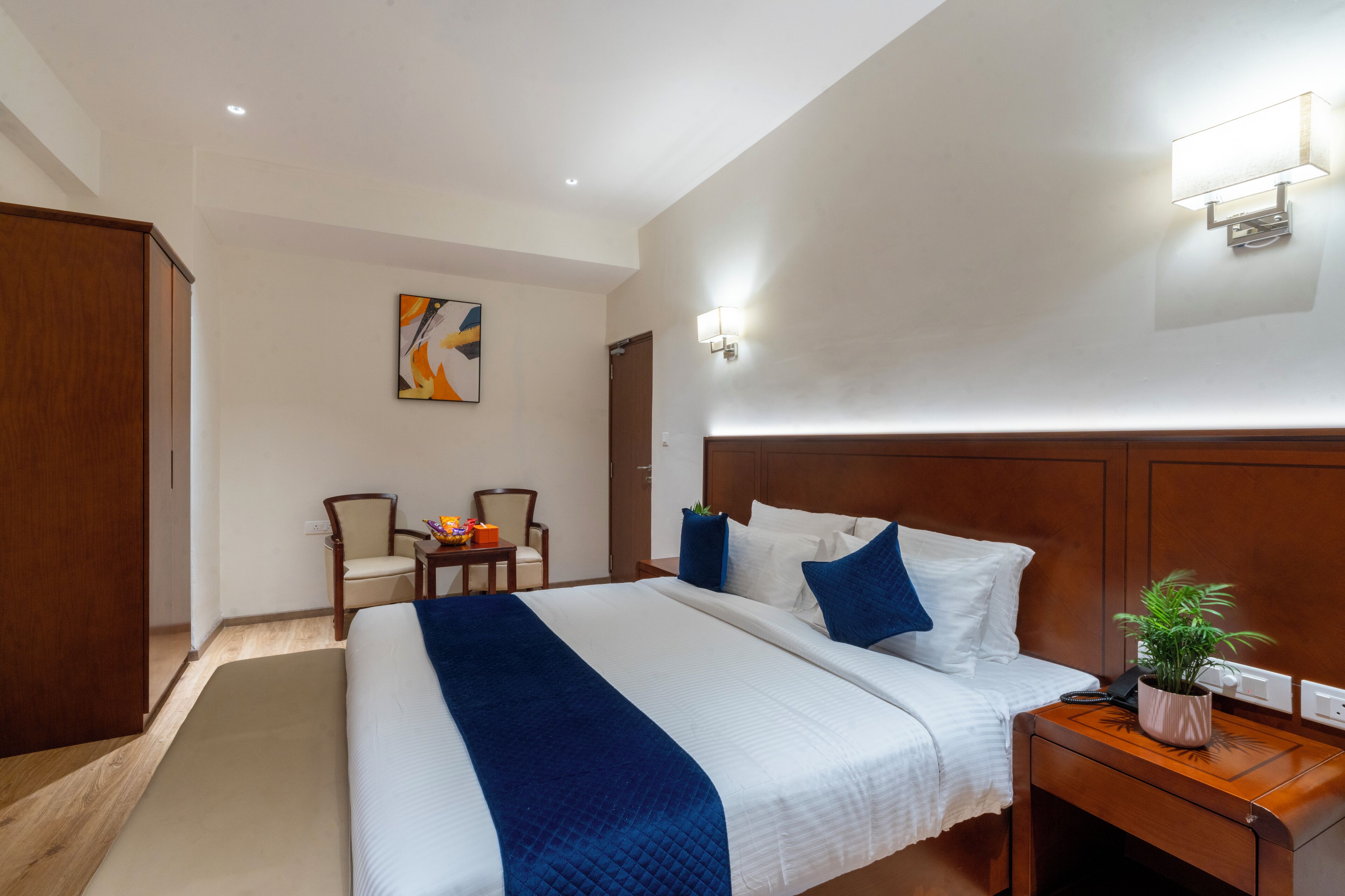 A hotel room with a large bed, a wooden headboard, a bedside table with a plant, and a desk with chairs.