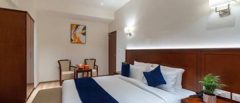 Quarto de hotel com uma cama grande, cabeceira de madeira, mesa de cabeceira com uma planta e uma escrivaninha com cadeiras.