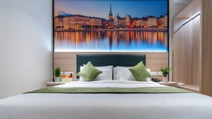 Ein Hotelzimmer mit einem großen Bett, grünen Kissen, einem Nachttisch mit Telefon und Pflanze sowie einem Stadtblick an der Wand.