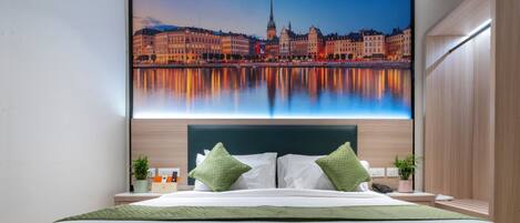 Habitación de hotel con una cama grande, almohadas verdes, una mesita de noche con un teléfono y una planta, y una vista de la ciudad en la pared.