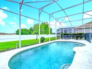 Une piscine dotée d’une zone abritée, entourée d’une clôture blanche, et un espace verdoyant avec des arbres en arrière-plan.