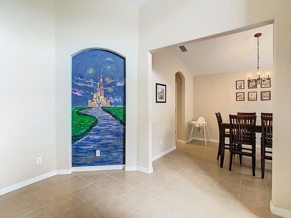 Un couloir orné d’une fresque représentant un château et des feux d’artifice, un coin repas avec un lustre, et une chaise blanche.