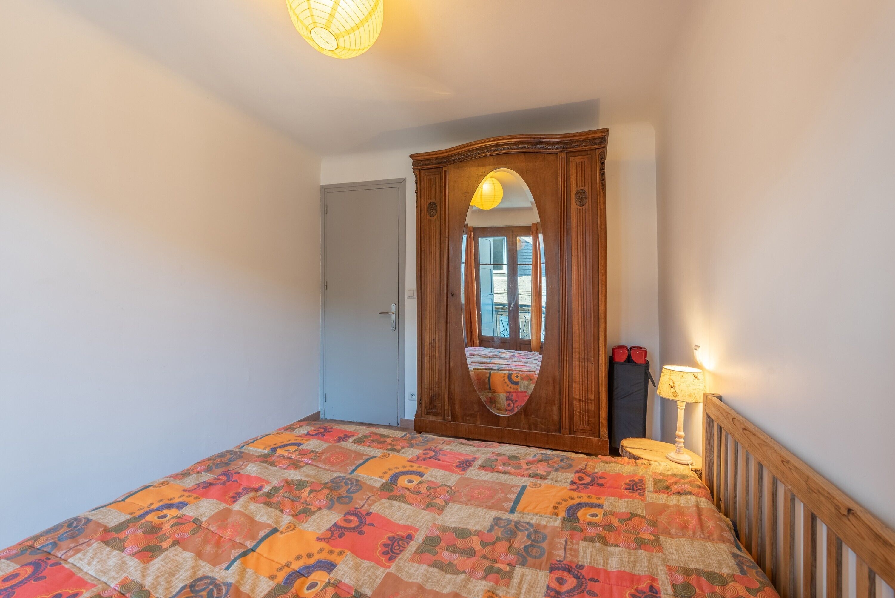 A bedroom with a bed, a wooden wardrobe, a mirror, a bedside lamp, and a door.