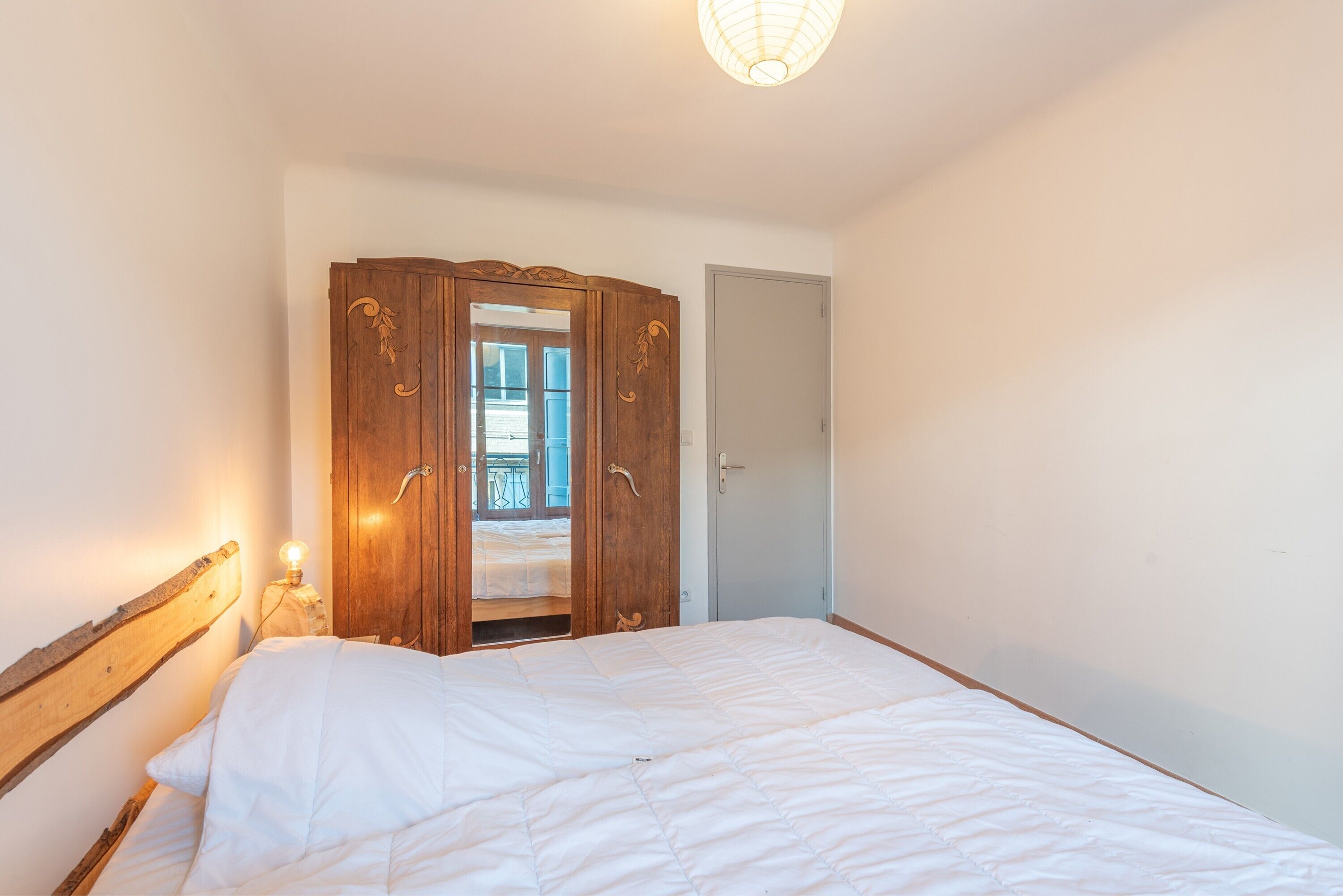 Et soveværelse med et træskab, en seng med hvide sengetøj, en senelampe og udsigt til en balkon gennem en dør.