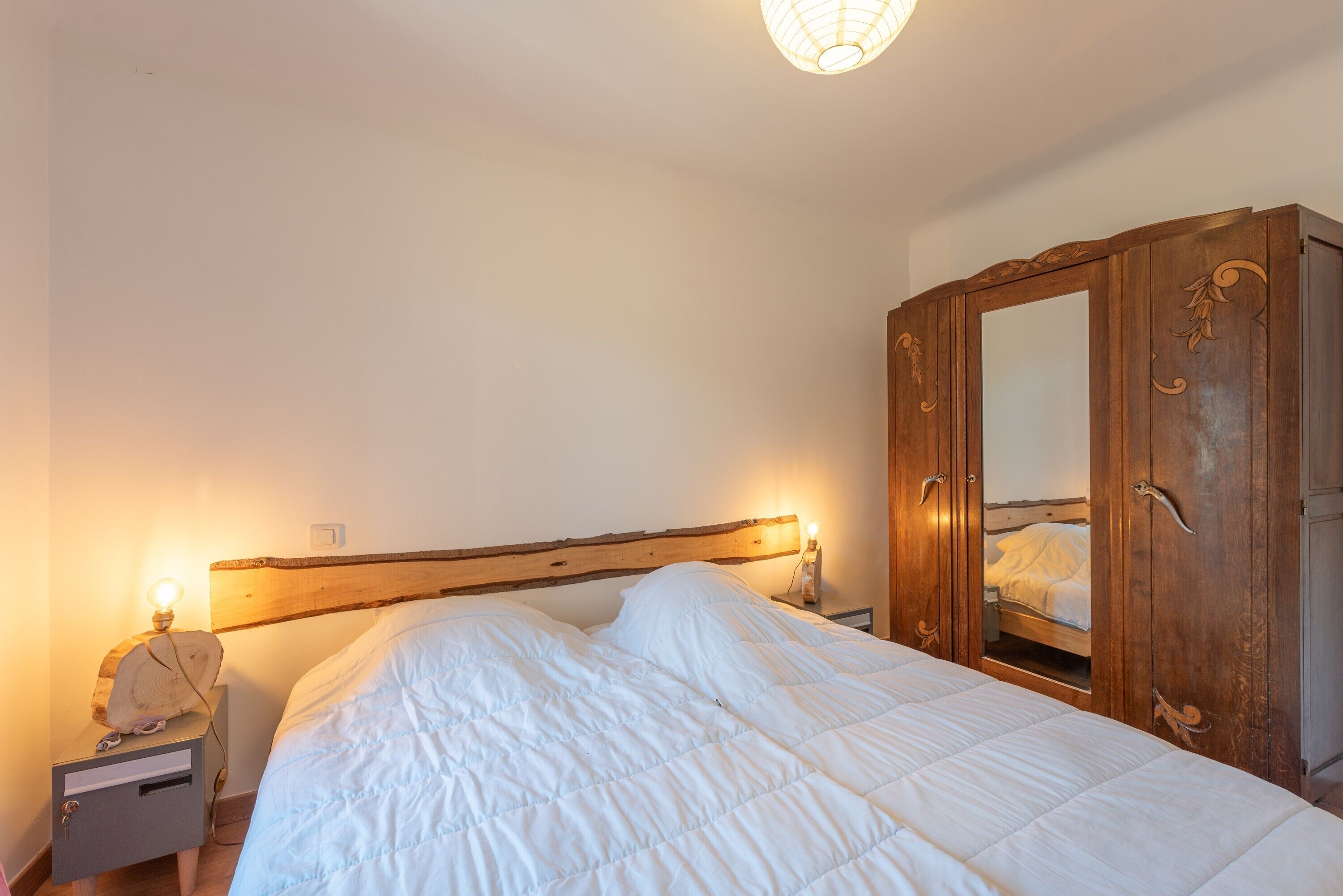 A bedroom with a wooden headboard, a bed with white bedding, a nightstand with a lamp, and a wardrobe with a mirror.