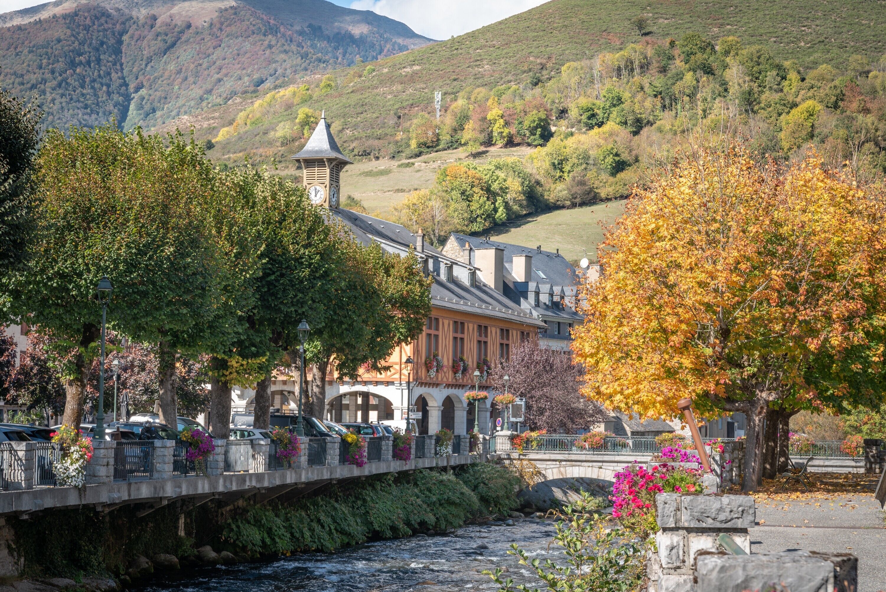 Un pont enjambant une rivière, avec un bâtiment doté d’un clocher, le tout entouré d’arbres et de feuillages d’automne.