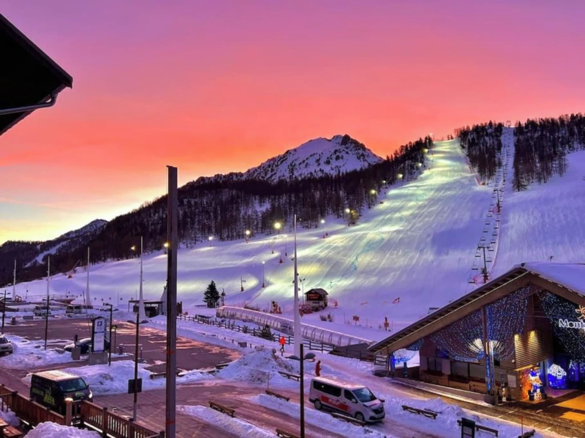 A ski resort with a mountain backdrop, a parking area, and a building with illuminated signage.