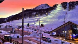 Estância de esqui com montanhas ao fundo, área de estacionamento e um edifício com letreiros iluminados.