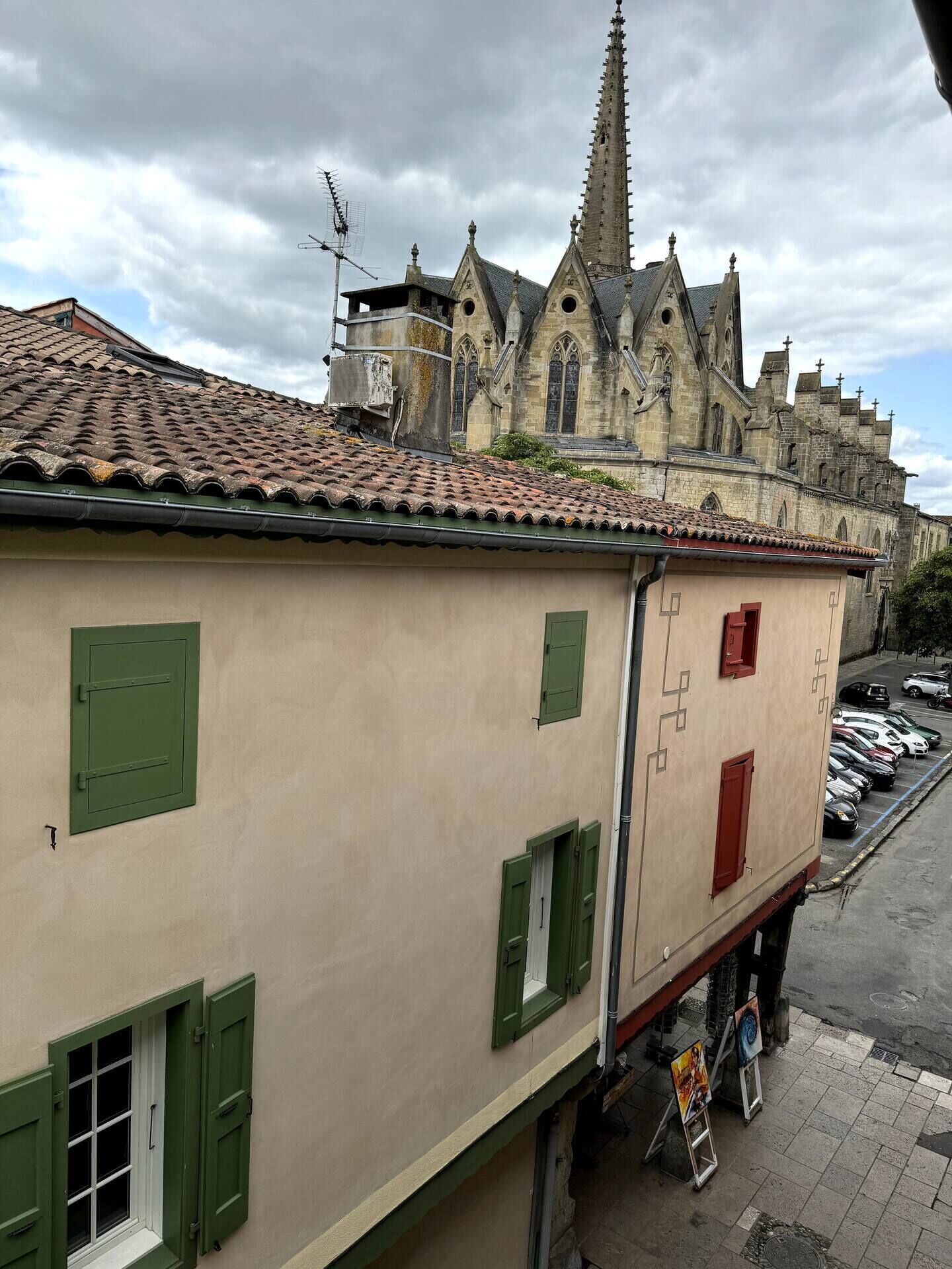 Un bâtiment avec un toit recouvert de tuiles, des volets verts et une église avec un clocher élancé en arrière-plan.