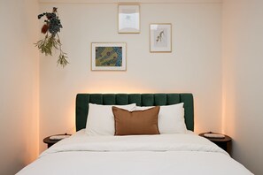 Un lit bien fait, doté d’une tête de lit recouverte d’un tissu vert, de draps blancs et d’un coussin décoratif marron. On y trouve deux tables de chevet avec des lampes, ainsi que trois œuvres d’art encadrées accrochées au mur.