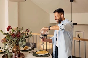 Une personne verse du vin dans un verre sur une table à manger, où se trouvent un vase de fleurs, des assiettes de nourriture et un intérieur moderne agrémenté d’une rambarde et d’un luminaire suspendu.