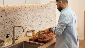 A person slicing bread on a wooden cutting board in a kitchen with a speckled backsplash and stainless steel sink. - The Brass Bear in Darlinghurst (Darlinghurst)