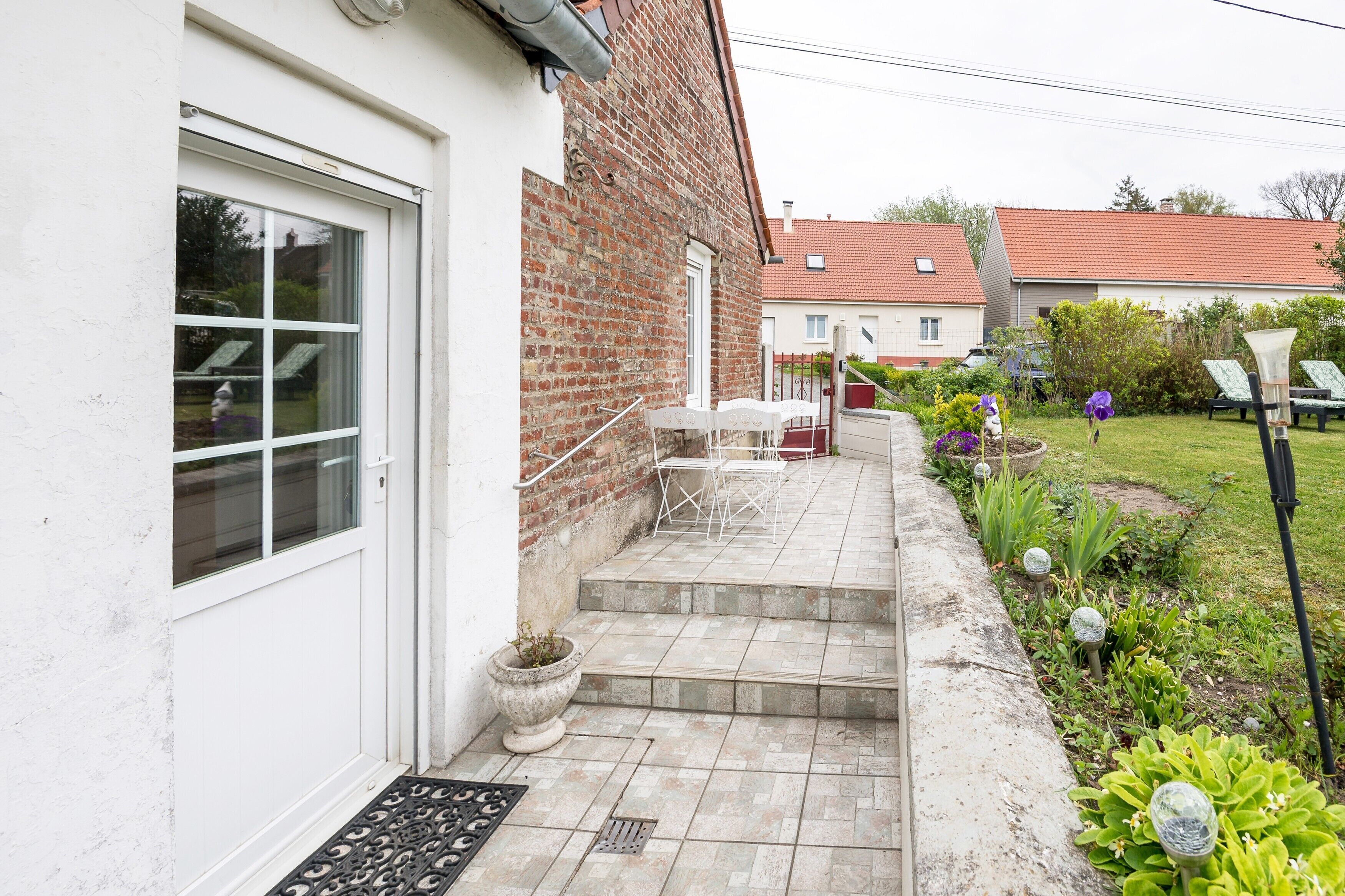 Une maison en briques avec une porte blanche, un petit patio avec une table et des chaises, et un jardin avec des plantes et un lampadaire.