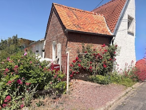 Une maison en briques avec un toit recouvert de tuiles rouges, une façade blanche et un jardin parsemé de fleurs roses.