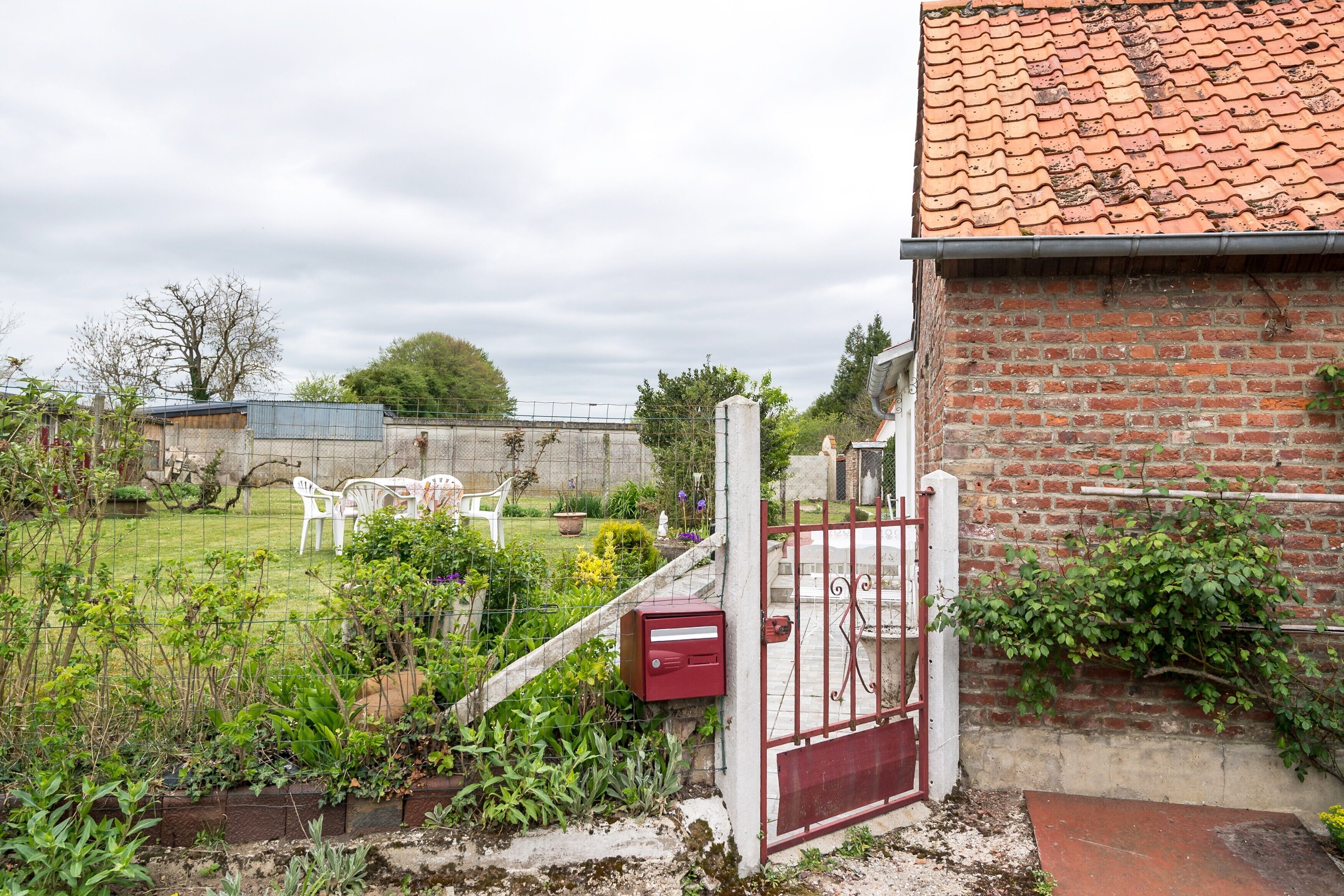 A fenced garden with a brick wall, a red gate, and a white chair.