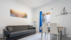 A modern living room with a grey sofa, white chairs, a dining table, a painting, and a sailboat decal on the wall. - Apartment 'De Playa' with Sea View, Balcony and Air Conditioning (Calafell)