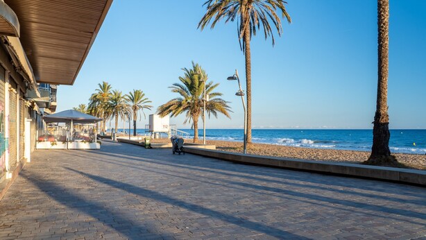 A paved walkway with palm trees, a beach, and a building with a sign '112' in the background. - Apartment 'De Playa' with Sea View, Balcony and Air Conditioning (Calafell)
