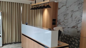 Una moderna recepción con detalles de madera y una encimera blanca, ubicada en un espacio con suelo de mármol y un amplio ventanal con cortinas.